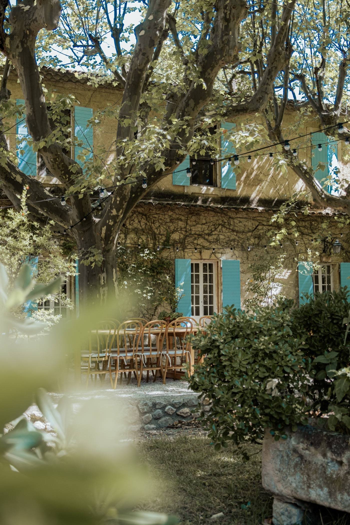 Bastide aux volets bleus et chaises en bois sous les arbres, au matin